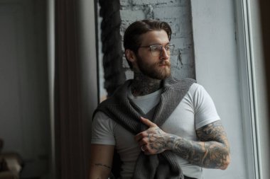 Moda gözlüklü, saç stili ve sakallı beyaz tişörtlü, süveterli yakışıklı hippi pencerenin yanındaki bir odada oturuyor.