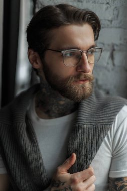 Sakallı, bıyıklı, beyaz tişörtlü, kapalı alanda örülmüş bir kazağı olan yakışıklı ve başarılı bir iş adamı. Hipster adamın portresi