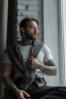 Süveterli, beyaz tişörtlü, dövmeli yakışıklı hippi, odanın penceresinde oturuyor.