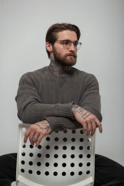 Moda örgü süveterli gözlüklü, saç stili ve sakallı havalı genç hippi stüdyoda beyaz bir sandalyede oturuyor.