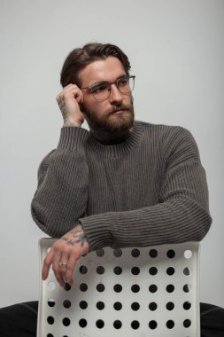 Moda örgü süveterli gözlüklü, saç stili ve sakallı havalı hippi yakışıklı adam stüdyoda beyaz bir sandalyede oturuyor.