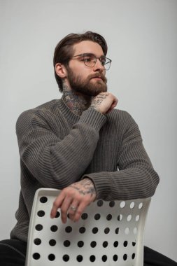 Moda gözlüklü, sakallı, bıyıklı, moda gözlüklü, şık bir genç adam stüdyoda beyaz bir sandalyede oturuyor.