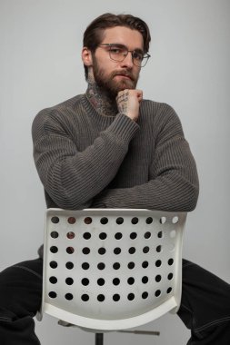 Hippi, yakışıklı, sakallı, saç stili, modaya uygun, gözlüklü, klasik süveterli bir adam stüdyoda beyaz bir sandalyede oturuyor ve poz veriyor.