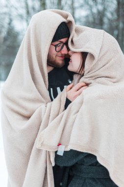 Sıcak bej bir battaniyeye sarılmış ve kış parkında kar altında birlikte duran mutlu genç aşıklar.