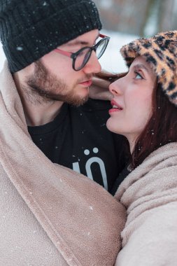 Güzel, genç bir çift, kış karlı bir parkta bej bir battaniyeyle sarılıyorlar. Aşk, sarılmalar ve temiz hava