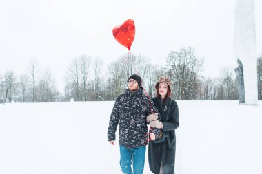 Şık, genç ve şık kışlık giysiler içinde kırmızı balonlu bir kalple beyaz bir parkta karla yürüyen güzel bir çift.