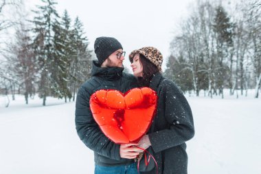 Kırmızı bir kalp balonu tutan ve kış parkında kar altında yürüyen şık kış kıyafetleri içinde mutlu ve genç aşıklar.