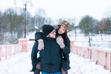 Moda kışı kıyafetleri içinde parkta eğlenen mutlu, neşeli aşıklar. Yakışıklı bir adam kız arkadaşını sırtında karın içinde taşır.
