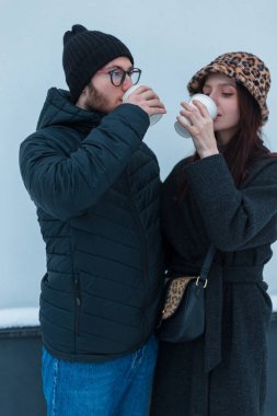 Genç yakışıklı bir adam ve şık kışlık giysiler içindeki güzel bir kadın, beyaz duvarın yanında duran bir çanta ve şapkayla kahve içiyorlar.