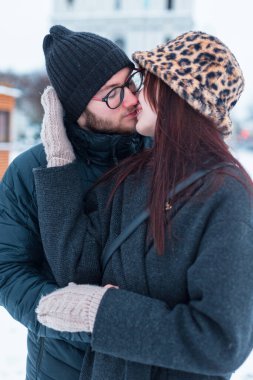 Güzel moda aşıkları, şık kışlık giysiler içinde, ceketli, şapkalı ve karlı şehirde sarılıp öpüşen çift.
