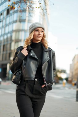 Şehirde siyah kazaklı, deri ceketli ve çantalı pantolonlu örgü şapkalı şık bir manken.