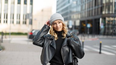 Güzel, modaya uygun, şapkalı, şık giyimli, deri ceketli ve çantalı genç bir kadın şehirde yürüyor.