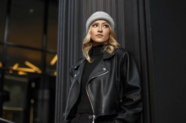 Moda desenli siyah deri ceketli ve süveterli, örgü örülü güzel bir kız modern siyah bir binanın yanında duruyor.