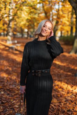 Altın bir sonbahar parkında çantalı, örgü örmüş, zarif bir elbisesi olan mutlu, güzel bir kadın.