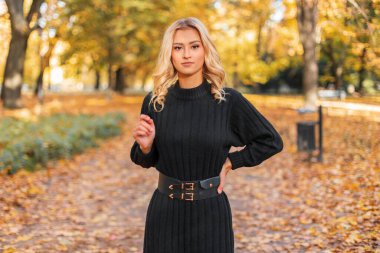 Güz parkında altın yeşilliklerle örülmüş siyah elbiseli güzel sarışın manken.