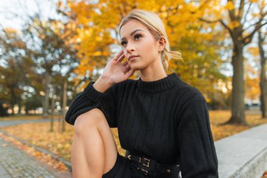 Şık, siyah örgü kazaklı güzel bir sonbahar kızı sonbahar parkında sarı yapraklarıyla oturuyor.