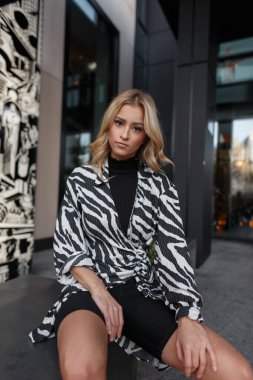 Şortlu şık bir zebra elbisesi giyen şık, sarışın bir kız şehirde oturuyor.