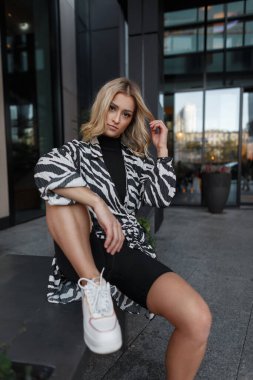 Havalı sokak güzeli, şık giyinen, zebra elbiseli, beyaz şortlu ve spor ayakkabılı sarışın kız binanın yakınında oturuyor.