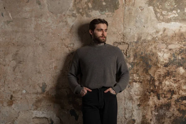 Sakallı, saç stili, desenli süveter ve beton bir duvarın yanında siyah kot pantolon giyen yakışıklı, vahşi bir adam.
