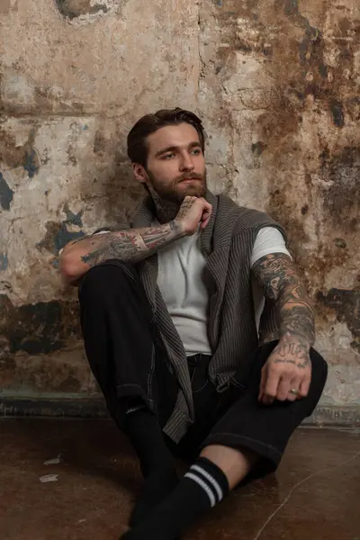 Süveteri ve siyah kotu olan beyaz tişörtlü, saç stili ve sakallı yakışıklı hippi, eski bir beton duvarın yanında oturuyor.