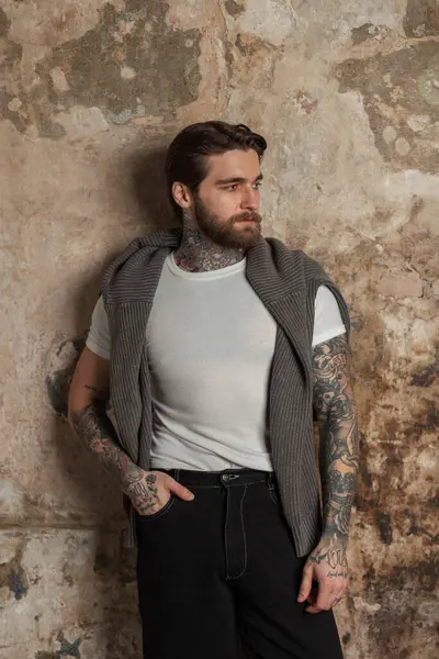 Sakallı, saç stili, beyaz tişörtlü, kazaklı ve kot pantolonlu yakışıklı hippi klasik beton duvarın yanında duruyor.
