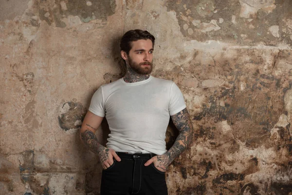 Havalı hippi vahşi bir adam, saç stili ve sakallı, modaya uygun beyaz tişörtlü, dövmeli eski moda bir grunge desenli duvarın yanında duruyor.