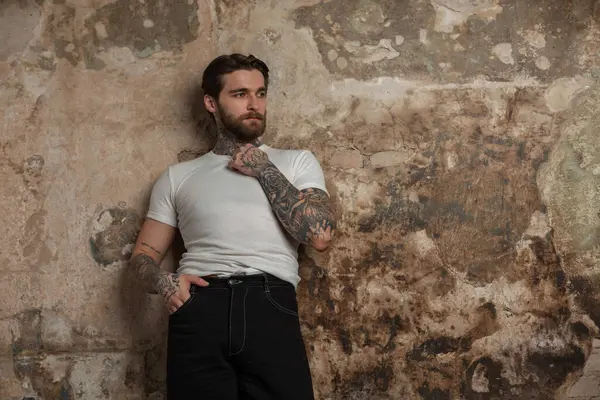 Süveterli beyaz tişörtlü, saç stili ve sakallı yakışıklı hippi eski beton bir duvarın yanında duruyor.