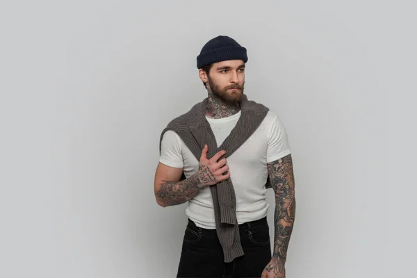 Cool hipster man with tattoo with hairstyle and beard in a fashionable white T-shirt and knitted sweater in the studio