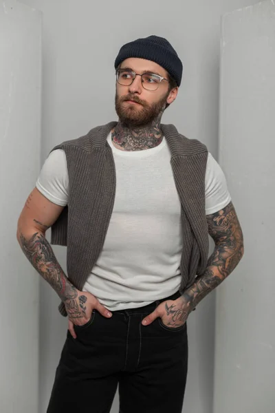 Hipster handsome cool man with a hairstyle and a beard with a tattoo with fashion glasses in a stylish white T-shirt and light stands on a white background in the studio
