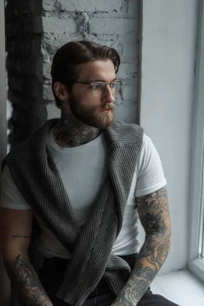 Sakallı, bıyıklı, saç stili, moda gözlüklü, beyaz tişörtlü, örgü kazaklı yakışıklı, genç hippi adam pencerenin yanında oturuyor.