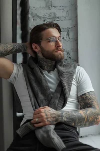 Sakallı, saç stili, dövmeli, beyaz tişörtlü gözlüklü, süveterli havalı hippi yakışıklı adam pencerenin yanında oturur ve rahatlar.