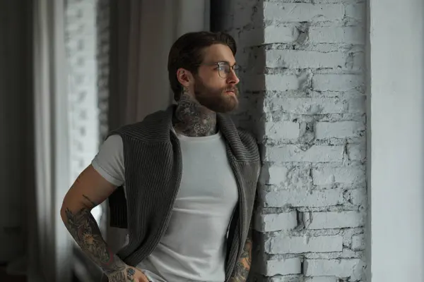 Successful handsome hipster man with tattoo and beard in fashion clothes with a white T-shirt and sweater stands near the window in the room