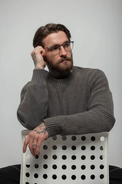 Moda örgü süveterli gözlüklü, saç stili ve sakallı havalı hippi yakışıklı adam stüdyoda beyaz bir sandalyede oturuyor.
