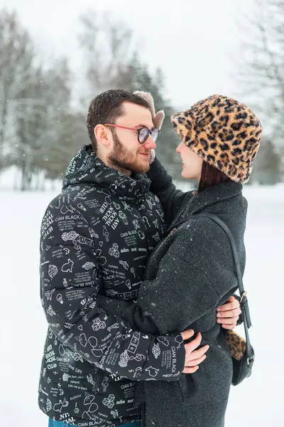 Güzel, mutlu, kış kıyafetleri içinde genç aşıklar karlı bir parkta durup sarılıyorlar.