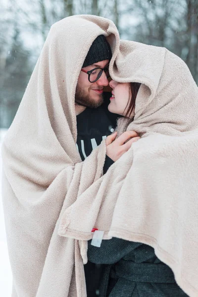Sıcak bej bir battaniyeye sarılmış ve kış parkında kar altında birlikte duran mutlu genç aşıklar.