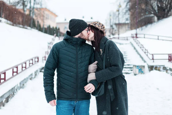 Kış kıyafetleri içinde güzel genç aşıklar karlı şehirde birlikte yürüyor ve öpüşüyorlar.