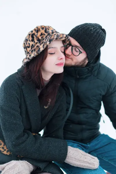 Güzel, genç, modaya uygun, kışlık elbiseli, şapkalı, ceketli ve eldivenli aşıklar bir kış günü dışarıda oturup sarılıyorlar.