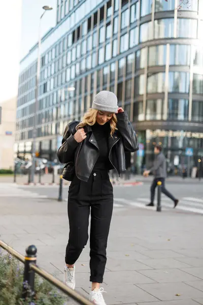 Şık giyinmiş, siyah kazaklı, deri ceketli, pantalonlu, beyaz spor ayakkabılı, şık bir kadın model şehirde dolaşıyor.