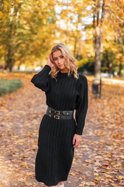 Güz parkında sarı yapraklı, siyah örgü elbiseli güzel sarışın bir kız yürüyor.