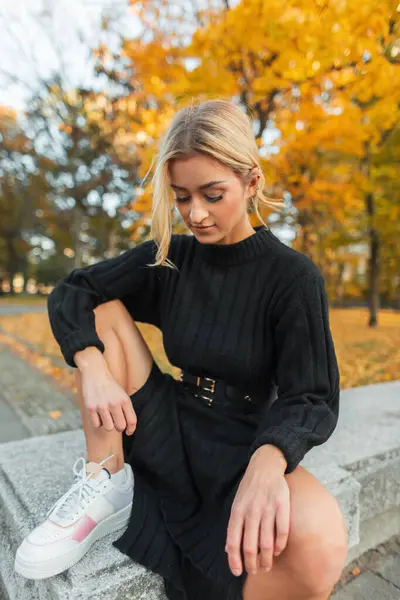 Güz parkında sarı yeşilliklerle oturup poz veren siyah elbiseli, beyaz spor ayakkabılı güzel sarışın manken kız.