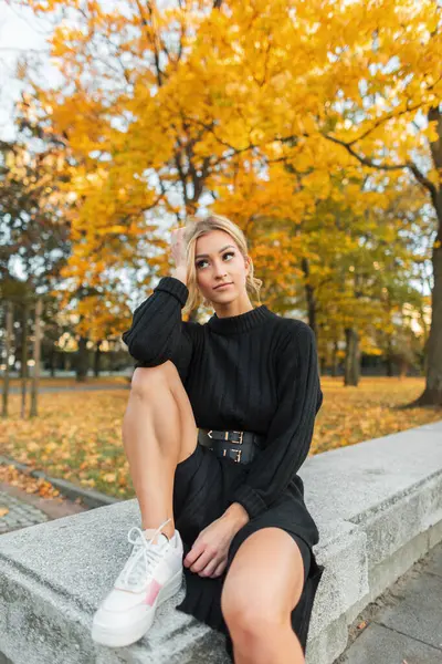 Moda elbisesi, seksi bacakları ve beyaz spor ayakkabıları olan şık bir kadın sarı bir parkta altın sonbahar yapraklarıyla oturuyor.