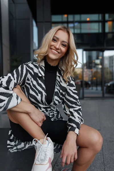 Şort ve spor ayakkabılarıyla şık bir zebra elbisesi içinde gülümseyen güzel sarışın kadın binanın yakınında oturuyor.