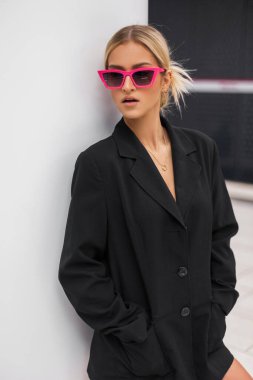 Havalı, güzel, alımlı, siyah stil bir spor ceketi olan, pembe güneş gözlüklü genç bir kadın beyaz bir duvarın yanında duruyor.