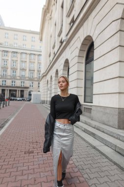 Şık, deri ceketli, siyah bluzlu ve etekli güzel bir kız şehirde yürüyor.