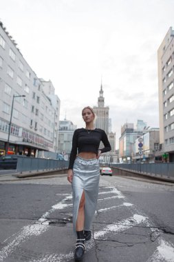 Moda kıyafetleri içinde, siyah bluzlu ve gümüş etekli güzel genç bir kadın şehirde yürüyor.