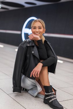 Mutlu moda şık elbiseli, deri ceketli güzel kız metro istasyonunda yerde oturuyor.