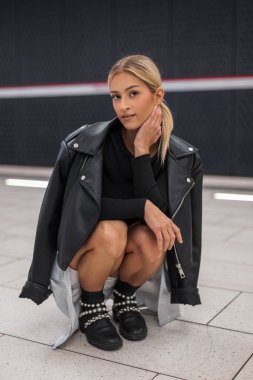 Moda kıyafetleri içinde, deri ceketli ve çizmeli etekli güzel bir kadın model metro istasyonunda yerde oturuyor.