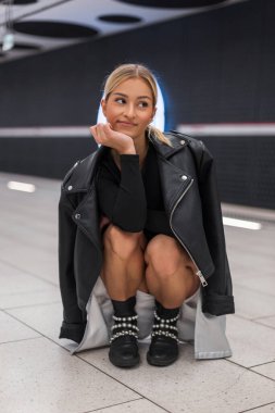 Deri ceketli, çizmeli etekli, şık giyinen güzel moda kadını modern bir metro istasyonunda oturur ve poz verir.