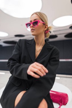 Siyah ceketli, pembe güneş gözlüklü havalı genç kız model metroda oturur ve poz verir.