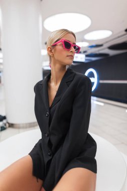 Pembe güneş gözlüklü, siyah ceketli, modern bir metro istasyonunda oturan ve poz veren genç ve çekici bir kadın.